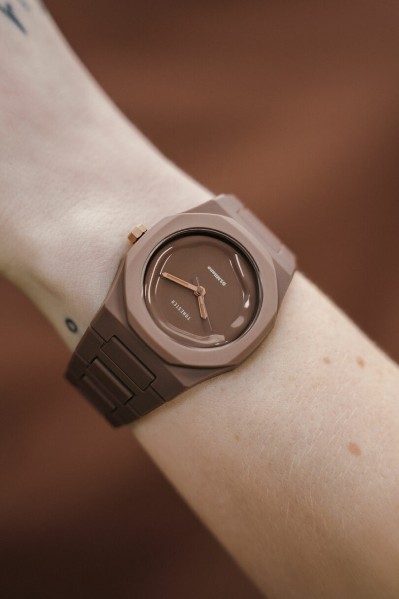 A close-up of a brown wristwatch with a matching strap on a person's wrist, featuring minimalist design and simple hour and minute hands.