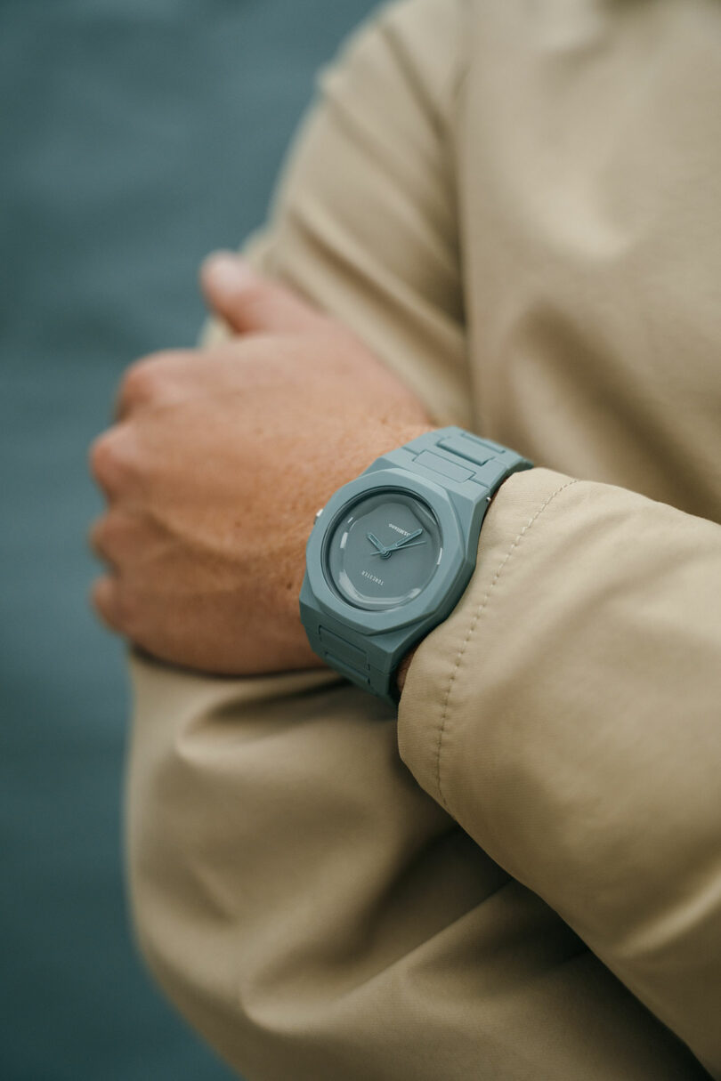 A person wearing a beige jacket has their arms crossed, displaying a matte green wristwatch with a minimalist design.