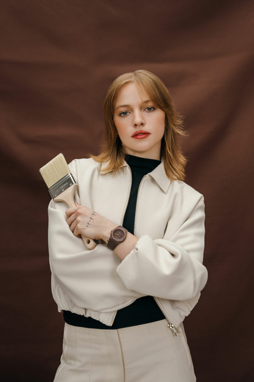 A woman with shoulder-length blonde hair wearing a cream-colored jacket holds a large paintbrush in one hand, standing in front of a brown backdrop.
