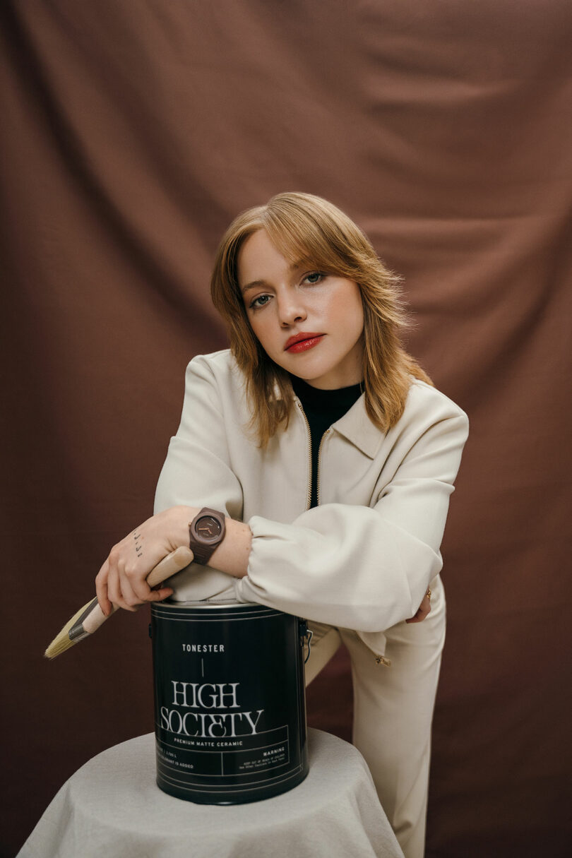 A woman in light-colored clothing leans on a black paint can labeled "High Society" and holds a paintbrush, with a brown backdrop behind her.
