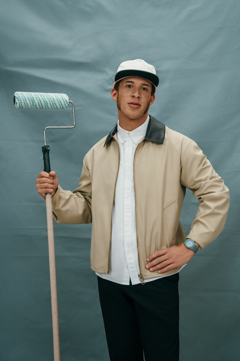 A person wearing a beige jacket, white shirt, black pants, and a cap holds a paint roller against a plain blue-gray background.