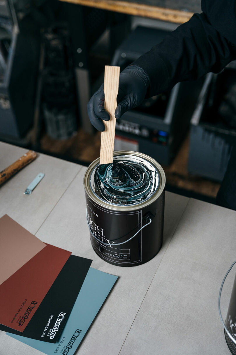 A person wearing a black glove stirs dark paint in a can with a wooden stick; paint color samples are laid on the table nearby.