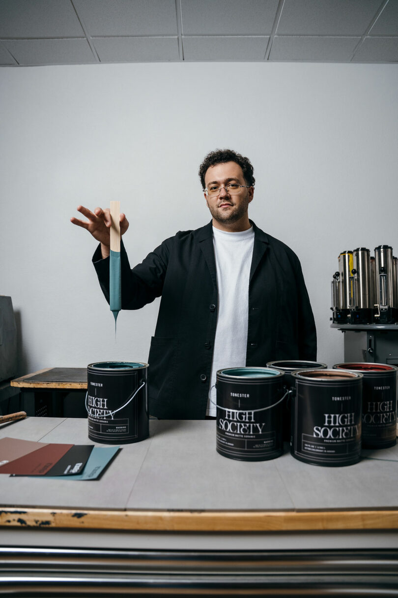 A person in a black coat dips a paint stick into a can labeled "HIGH SOCIETY" on a table with other paint cans and color samples in a studio setting.