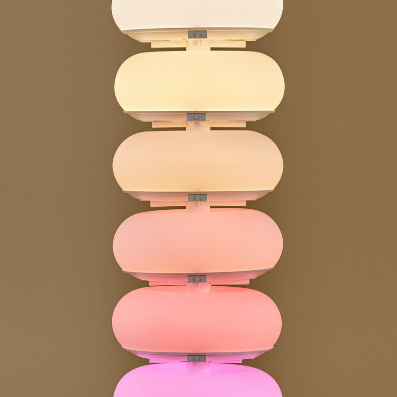 A vertical stack of rounded, translucent light fixtures in gradient colors from cream at the top to pink at the bottom, mounted on a brown wall.