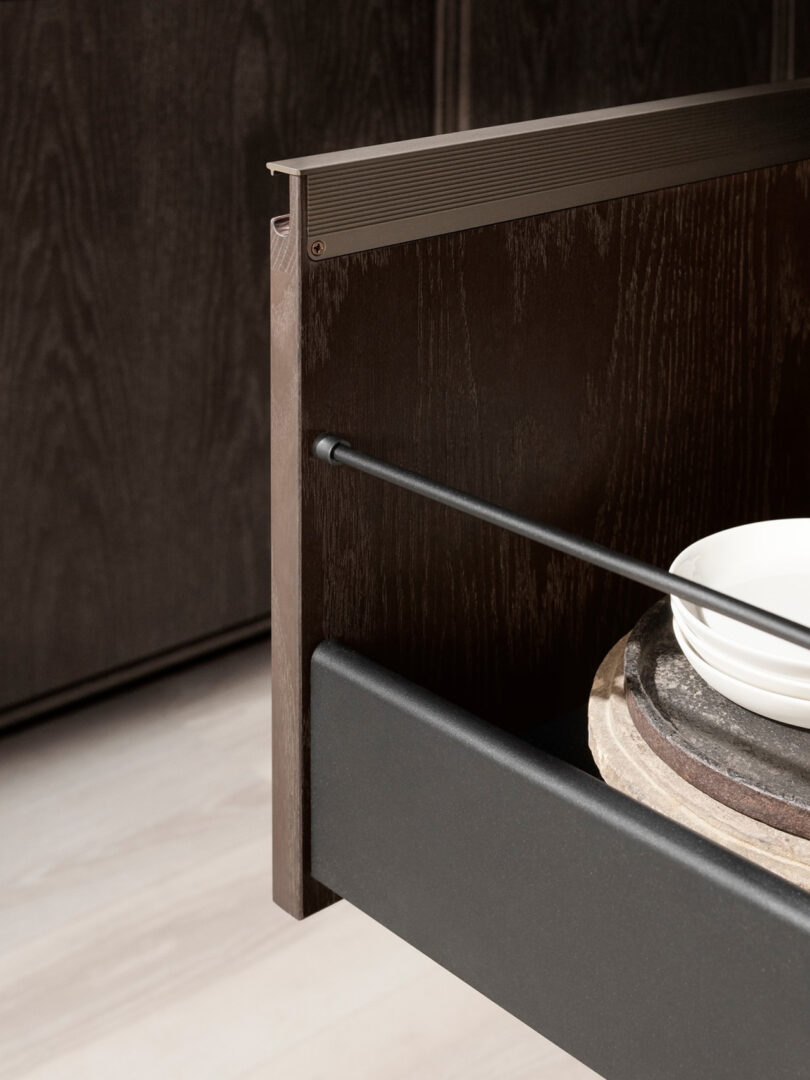 Open Vipp kitchen drawer with a dark wood finish, containing a round wooden board and a white bowl, set against a sleek dark cabinet background.