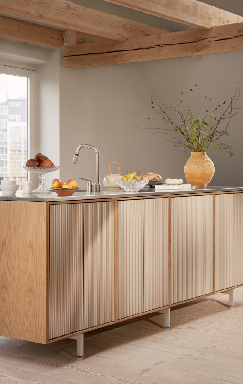 Modern Vipp kitchen with light wood cabinets, a large vase with branches, assorted baked goods, fruit, and a window revealing a cityscape in the background.