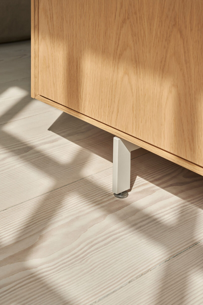 Close-up of a Vipp light wood cabinet corner with a metal leg, casting shadows on a light-colored wooden floor.