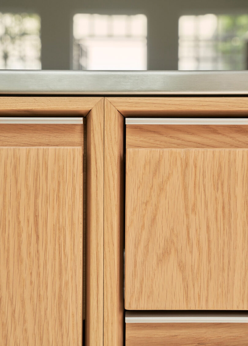 Close-up view of light wood Vipp kitchen cabinets with a metal countertop, set against a blurred background with bright windows.