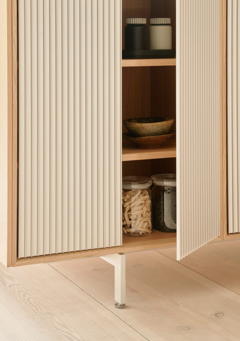 A Vipp wooden cabinet with grooved white doors is partially open, revealing shelves with jars, bowls, and containers inside. The cabinet stands on a light wood floor.