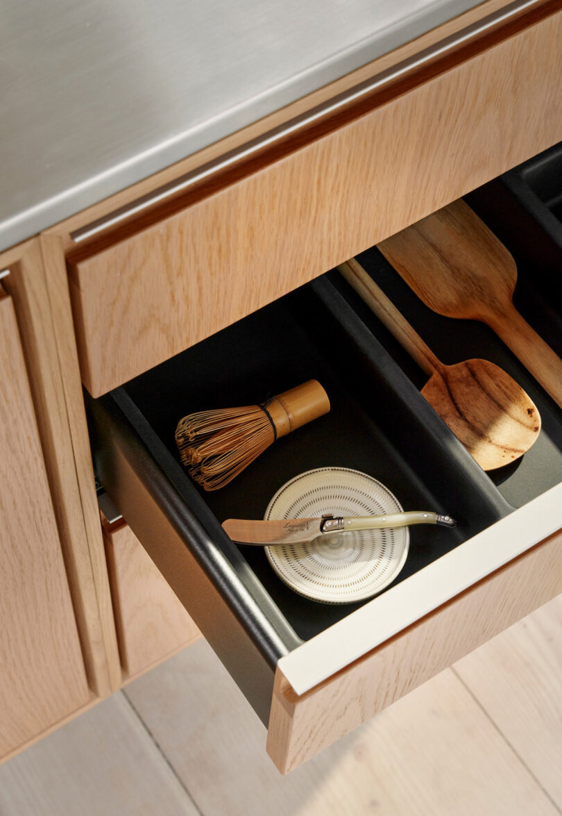An open Vipp kitchen drawer contains a bamboo whisk, a metal strainer, a wooden spoon, and a wooden spatula, all neatly arranged under a sleek metal countertop.
