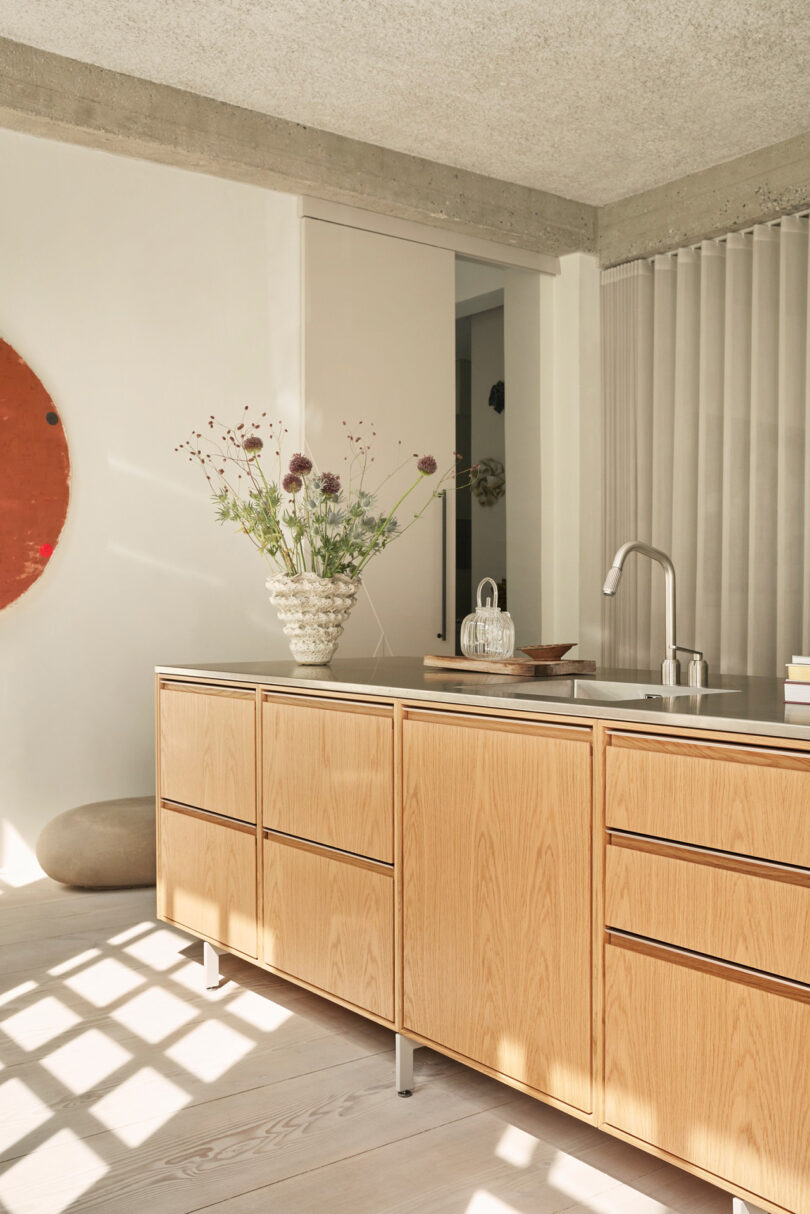 A modern kitchen with light wood cabinets, a sleek Vipp metal countertop, a vase of flowers, and minimal decor, illuminated by sunlight through a window.