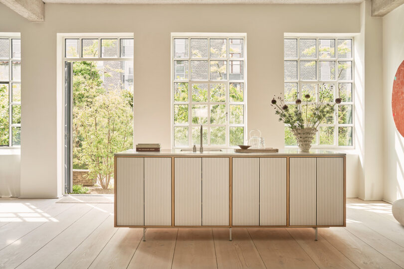 Minimalist Vipp kitchen with a white island, large windows letting in natural light, and a vase with flowers on the counter; garden visible outside.