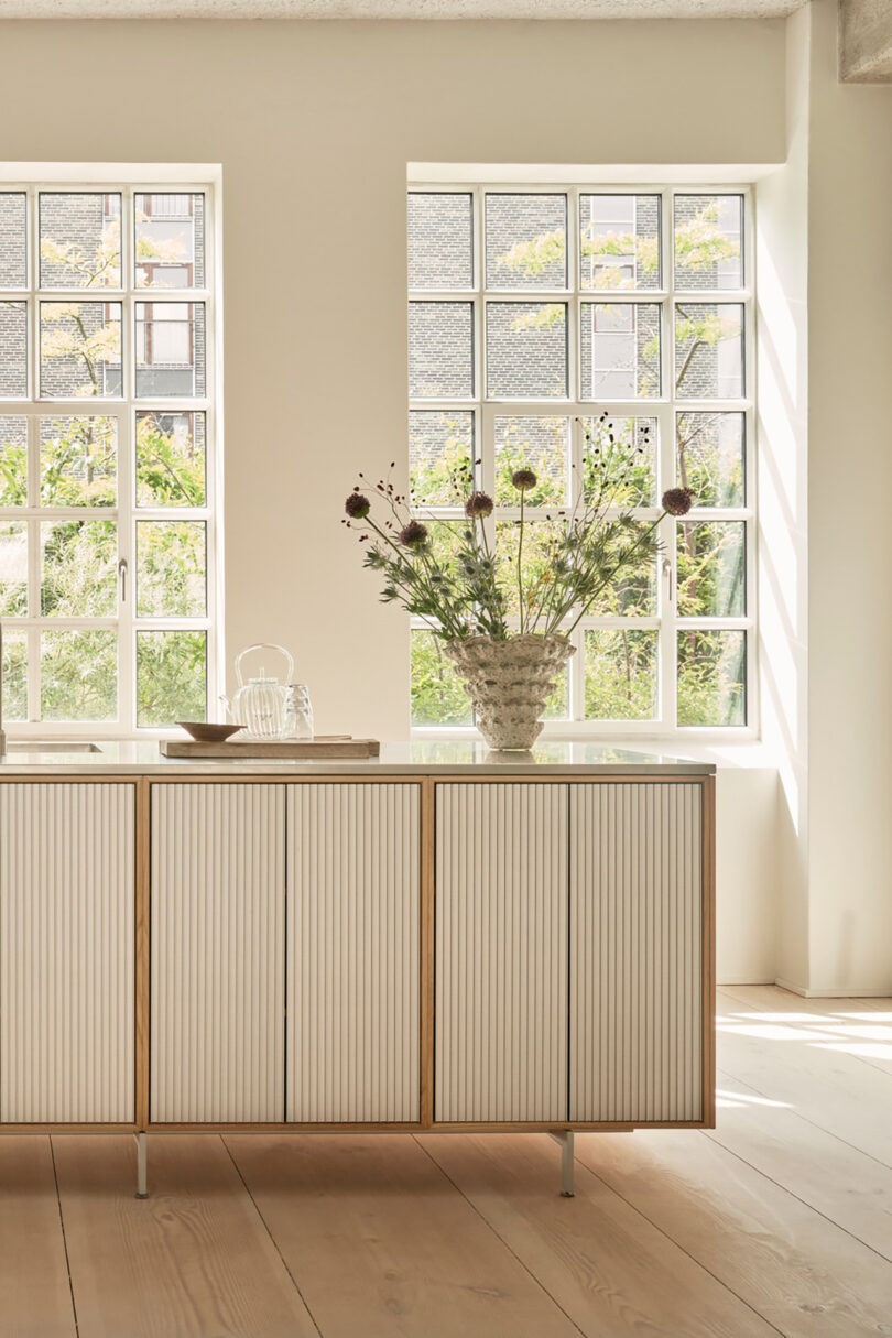 A light-filled room with large windows, a Vipp wooden sideboard, a vase with wildflowers, and a glass teapot on a tray.