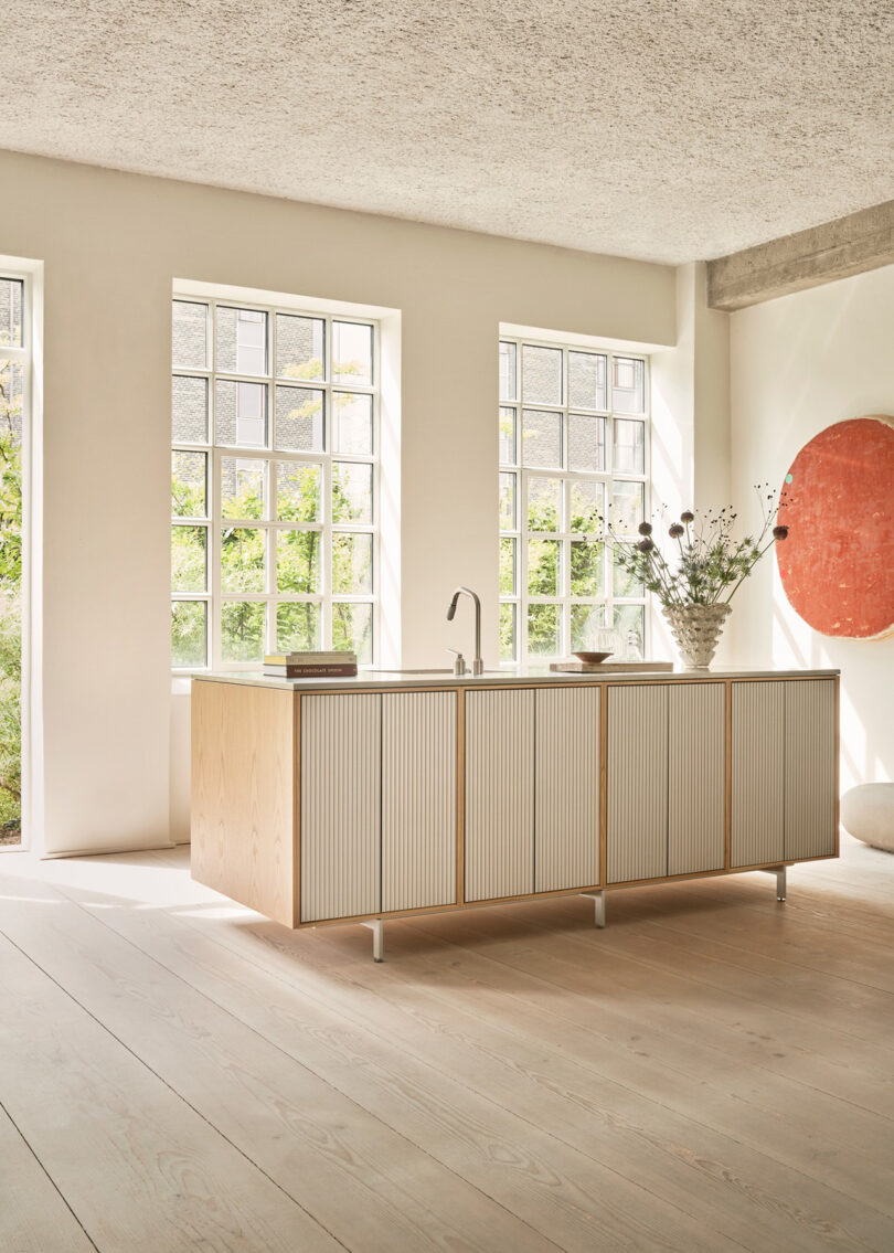 Minimalist Vipp kitchen with a central island, large windows, light wood flooring, a vase of flowers, and a striking red circular wall art piece.