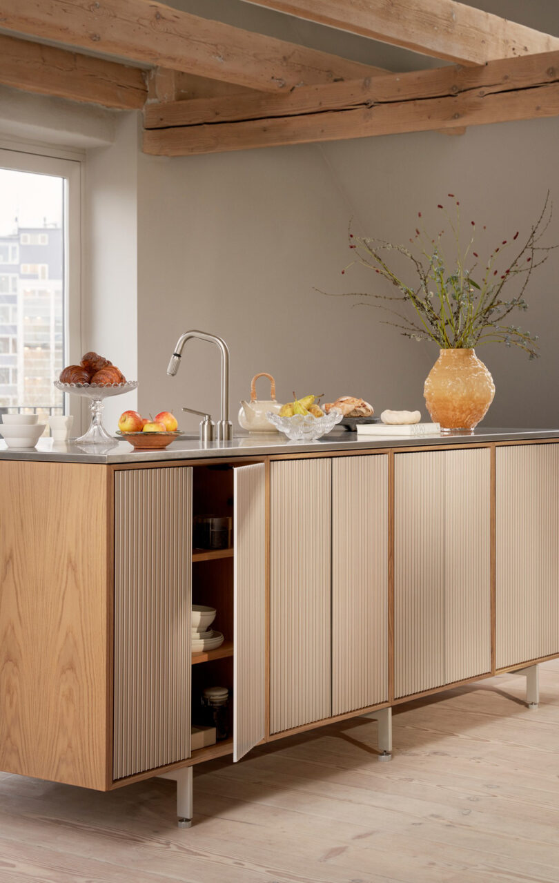 A modern kitchen island with Vipp wood paneling, an open cabinet, fruit, pastries, a vase with branches, and a city view through the window.