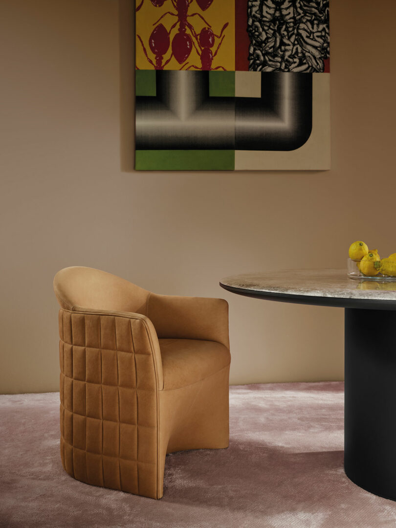 A tan Wittmann upholstered chair with a quilted back sits next to a round marble table; abstract artwork hangs on a beige wall above mauve carpet. Lemons are on the table.