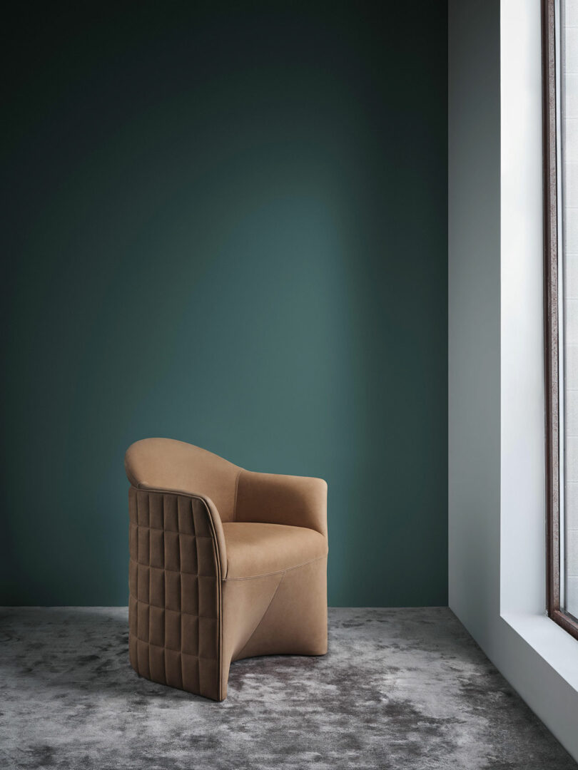 A tan, quilted Wittmann armchair sits on a gray carpet in a minimalist room with a dark green wall and a large window letting in natural light.
