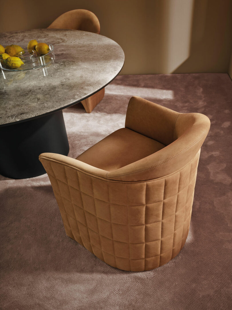 A Wittmann tan upholstered chair with a quilted backrest sits next to a round marble table with a glass bowl of lemons, on a soft mauve carpet in a modern interior setting.