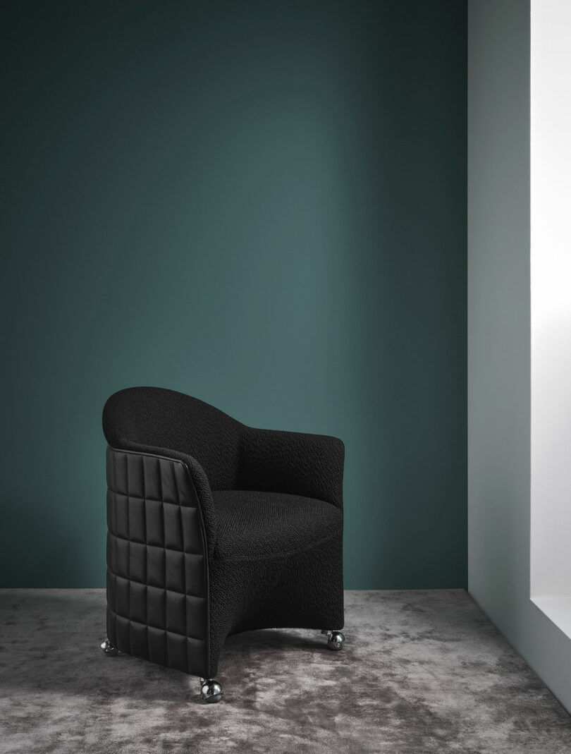 A modern black Wittmann armchair with a quilted side design sits on a gray carpet against a dark green wall in a minimalist room with natural light.