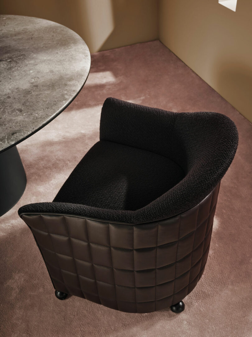 A black textured Wittmann armchair with a quilted leather exterior sits on a mauve carpet next to a round stone table in a modern room with beige walls.